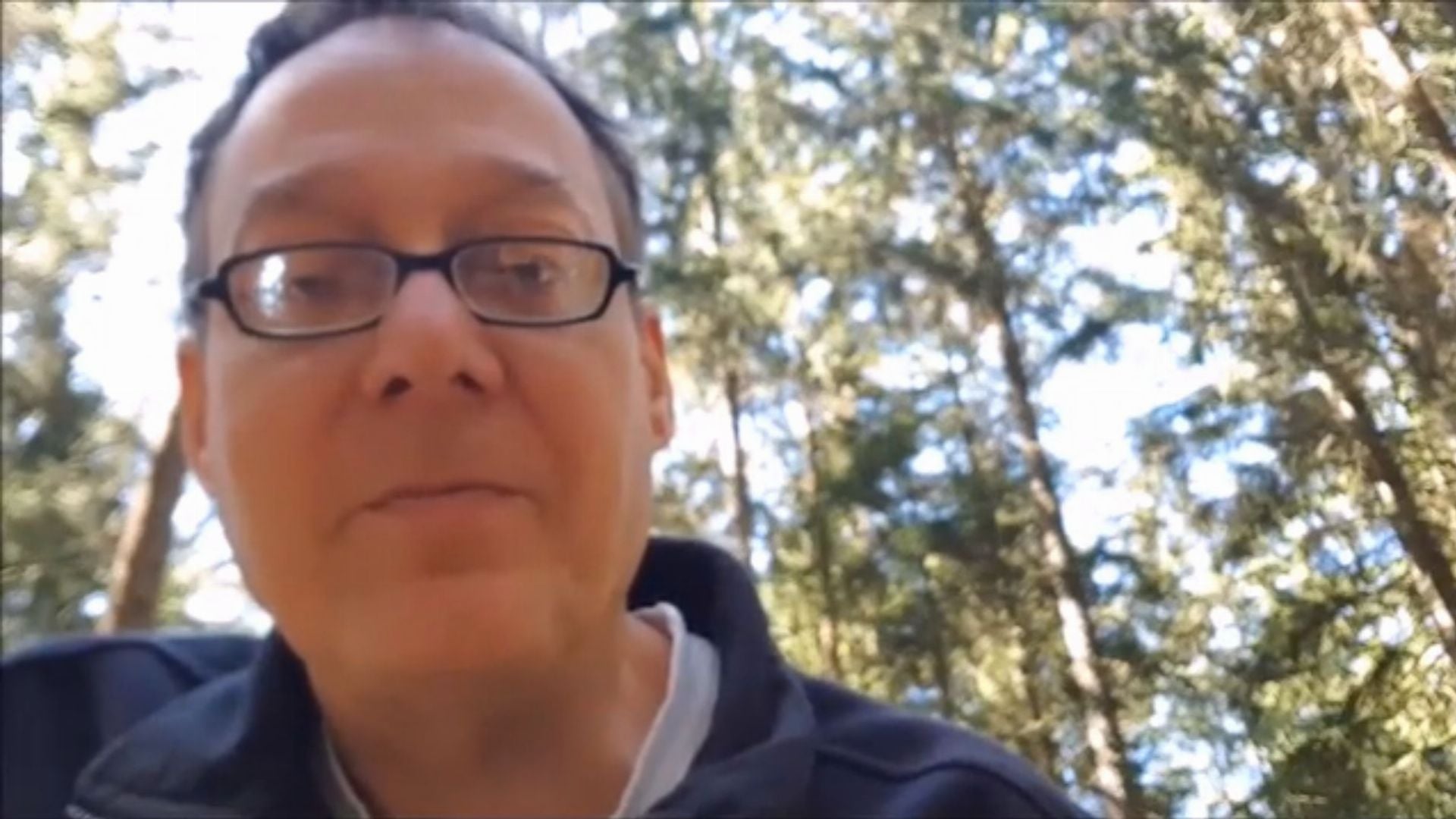11. Für die Ernte bereit sein. Middle-aged man with glasses, wearing a black jacket over a gray hoodie, making a slight smile against a backdrop of tall, dense trees under a clear sky. Opens a video in a new window.