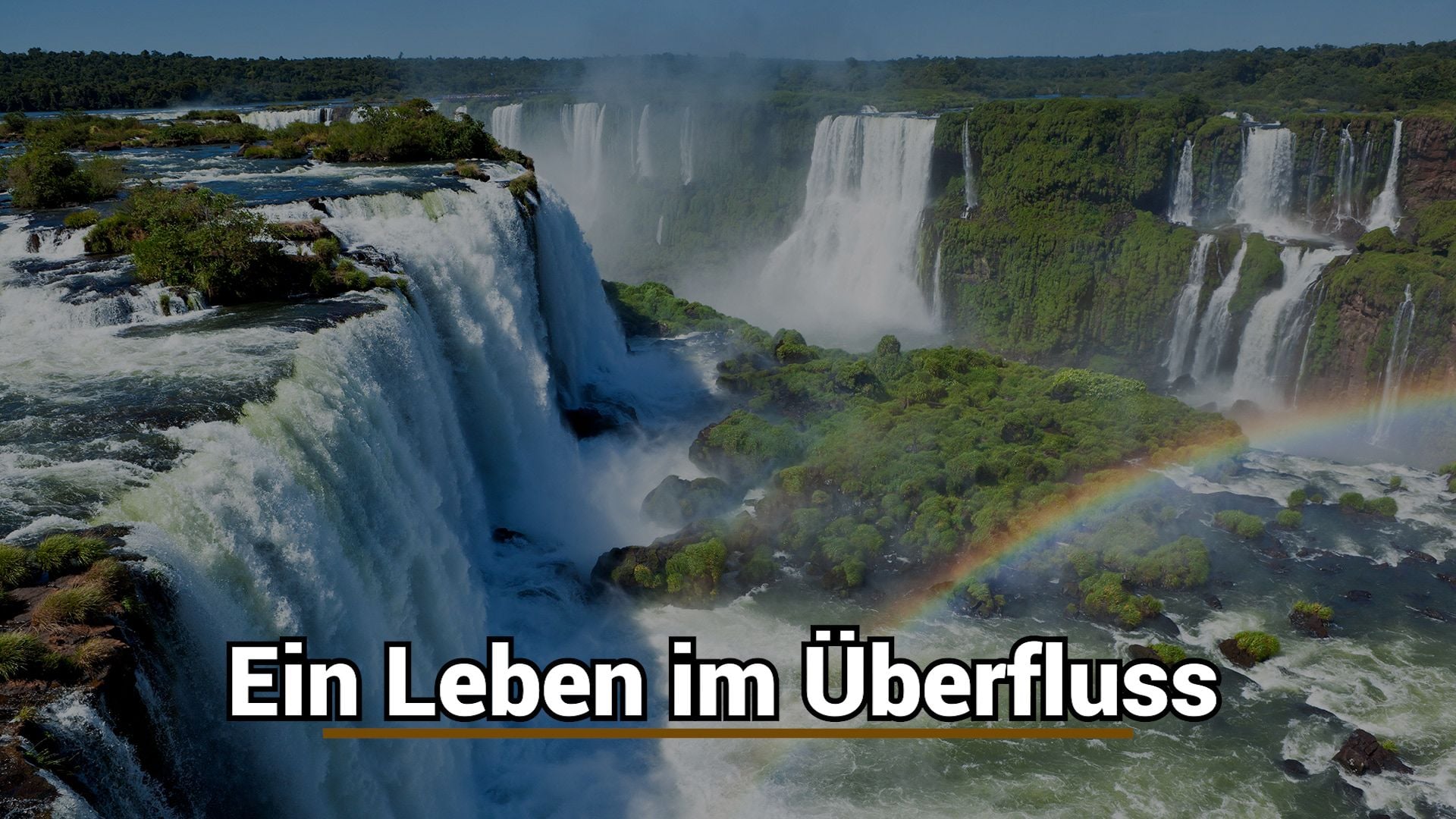 6. Ein Leben im Überfluss. A sweeping aerial view of a majestic waterfall system flanked by lush greenery and a vibrant rainbow arching across the mist-filled air. Text overlay in German reads "Ein Leben im Überfluss." Opens a video in a new window.