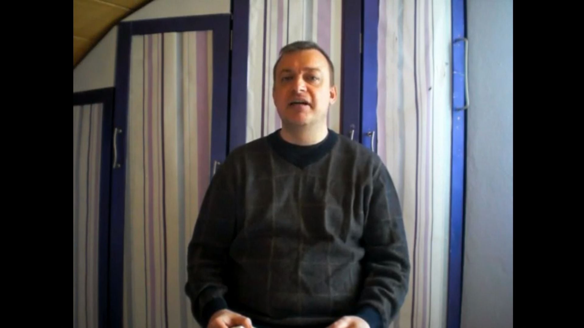 2. Zeichen am Himmel. A man in a dark sweater stands speaking, with striped curtains in various shades of blue and purple behind him. His expression is earnest. Opens a video in a new window.