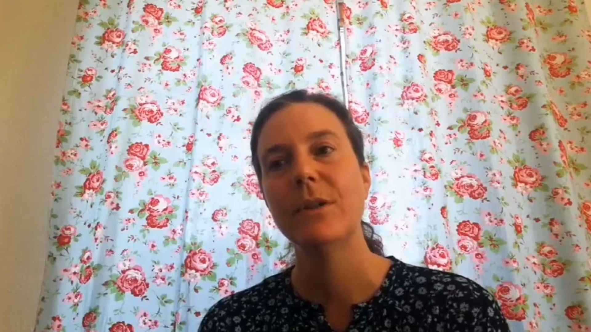 12. Sehnsuchtsbotschaft für Dich. A woman with a calm expression is visible from the chest up, wearing a dark blue top with subtle patterns. She is set against a backdrop of a curtain featuring a floral pattern with pink roses on a light background. Opens a video in a new window.