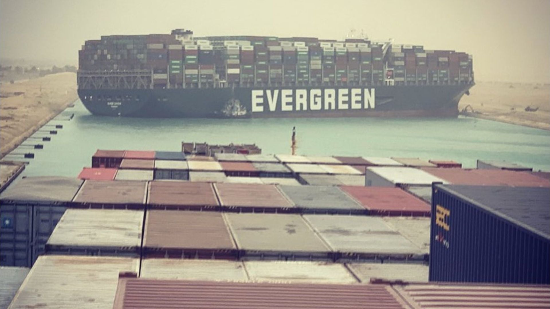 4. Salid, Parte I – Los cuatro vientos están soplando. A large cargo ship named EVERGREEN is positioned diagonally across a narrow canal, surrounded by sandy banks under a hazy sky. In the foreground, numerous colorful shipping containers are stacked at a port. Opens a video in a new window.