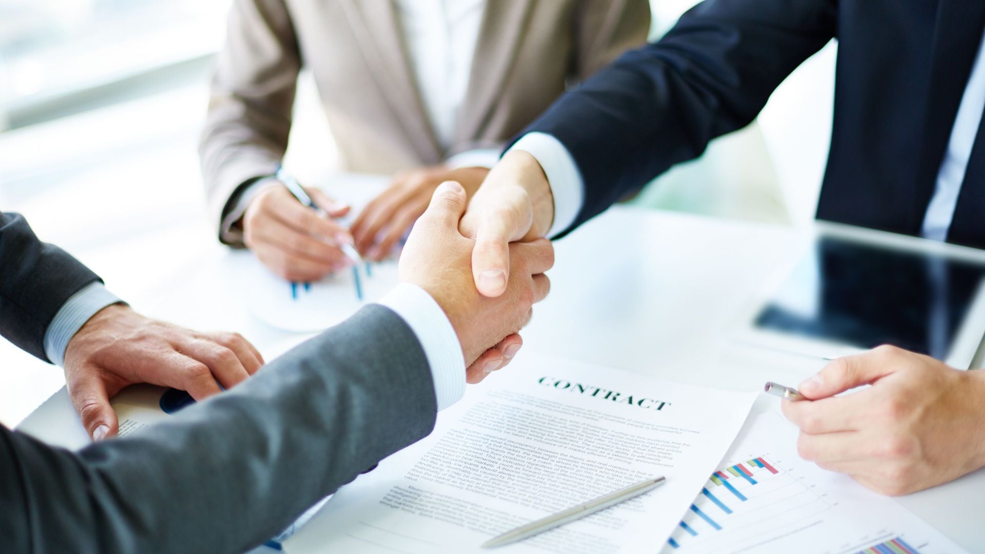 6. Was ist der Ewige Bund?. A close-up photo showing two individuals engaging in a firm handshake over a table with business documents and financial charts spread out, implying the closure of a successful negotiation. Another person appears to be taking notes, participating in the agreement process. Opens a video in a new window.