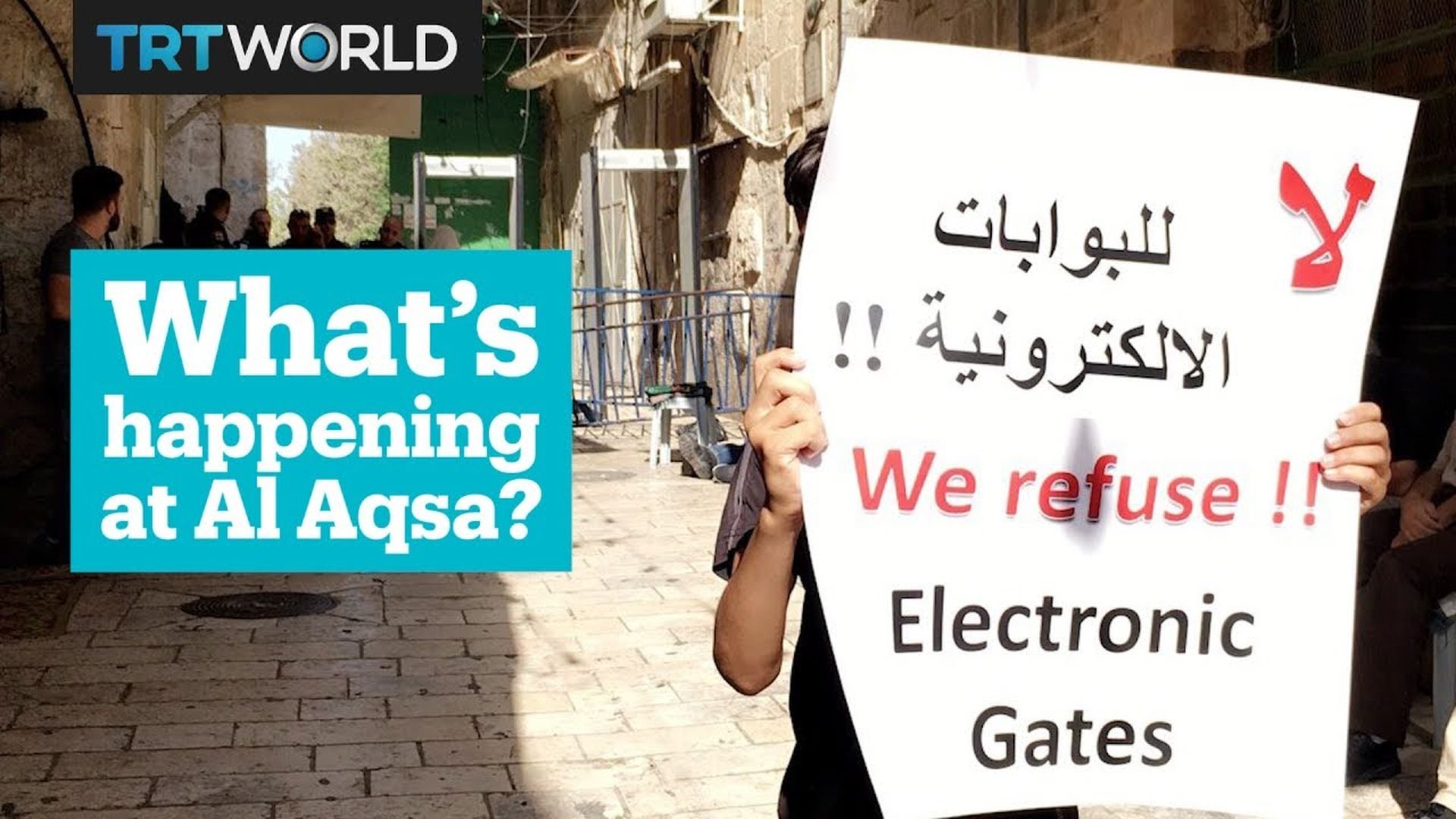 2. Al Aqsa—It's Not About Security. A person in a street holding two signs, one in English and one in Arabic. The English sign reads, "We refuse!! Electronic Gates." To the left, a graphic element with the text "What’s happening at Al Aqsa?" is visible. The scene features Middle Eastern architecture in the background, conveying a public demonstration or protest atmosphere. Opens a video in a new window.