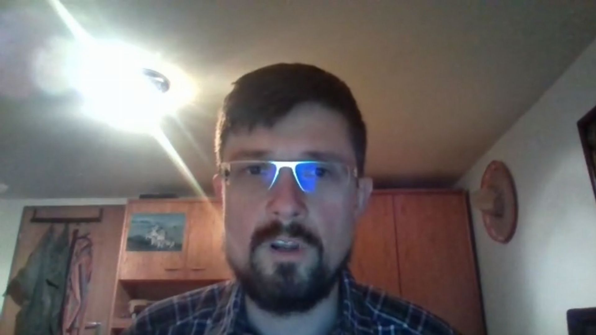 7. Die Wiederkunft unseres geliebten Herrn Jesus Christus Alnitak Powehi. A man with a beard and glasses sits indoors with an illuminated light source reflecting off his glasses, casting a bright flare on the left side of the image. A wooden cabinet and a wall hanging are visible in the dimly lit background. Opens a video in a new window.