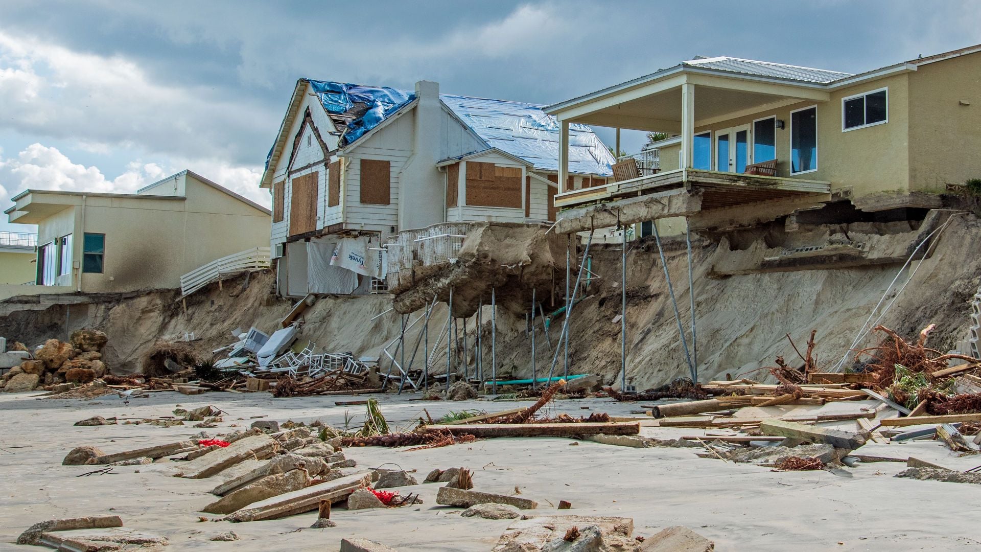 3. Construido sobre la arena. Coastal erosion has caused significant damage to seaside homes, leaving structures precariously supported on eroded cliff sides with visible debris scattered across the beach. Opens a video in a new window.