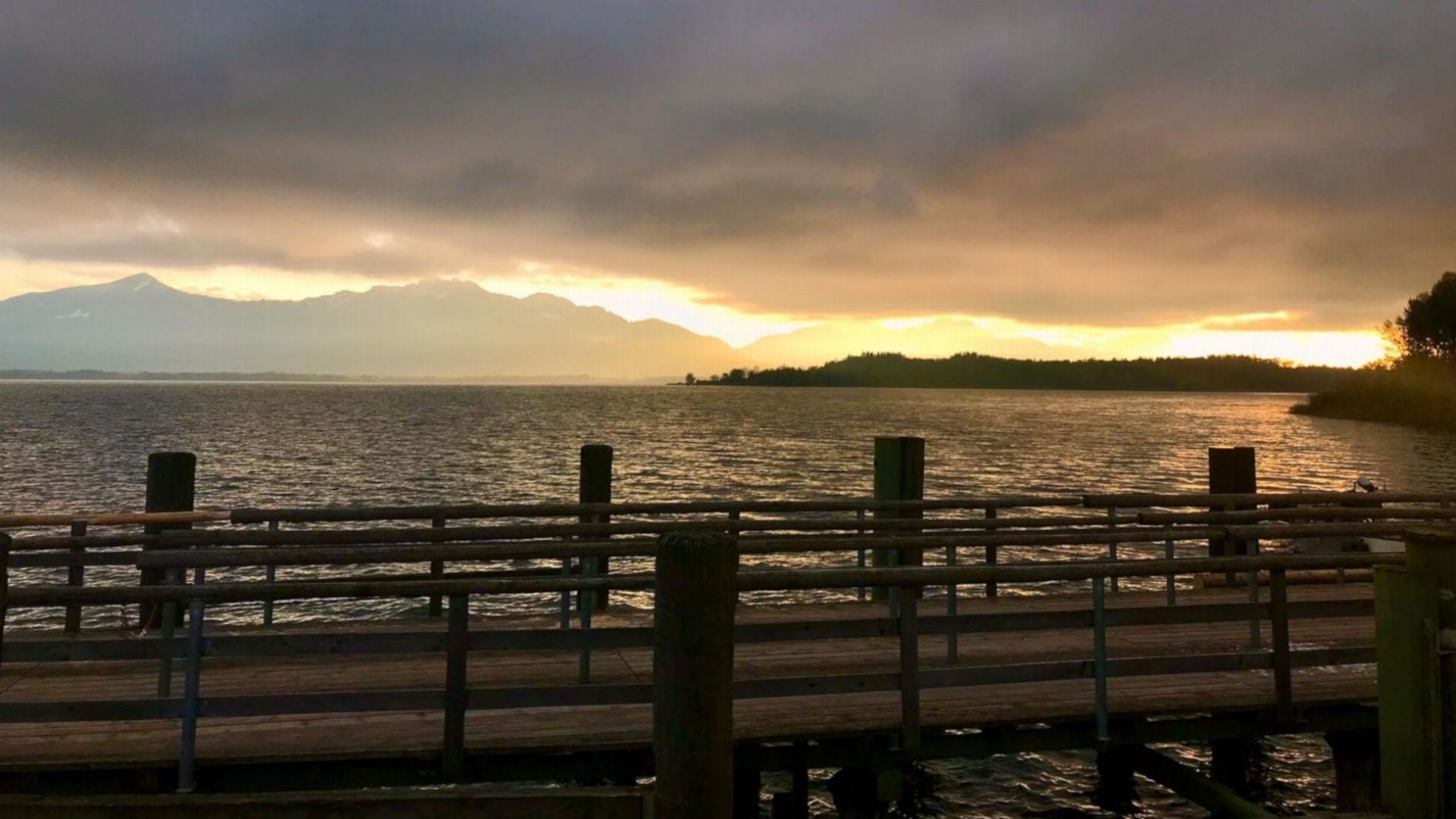 25. 144 Luftballons. A serene sunset view over a large body of water, featuring silhouettes of distant mountains under a dramatic sky. The foreground shows a wooden pier with several posts, enhancing the panoramic nature scene. Opens a video in a new window.