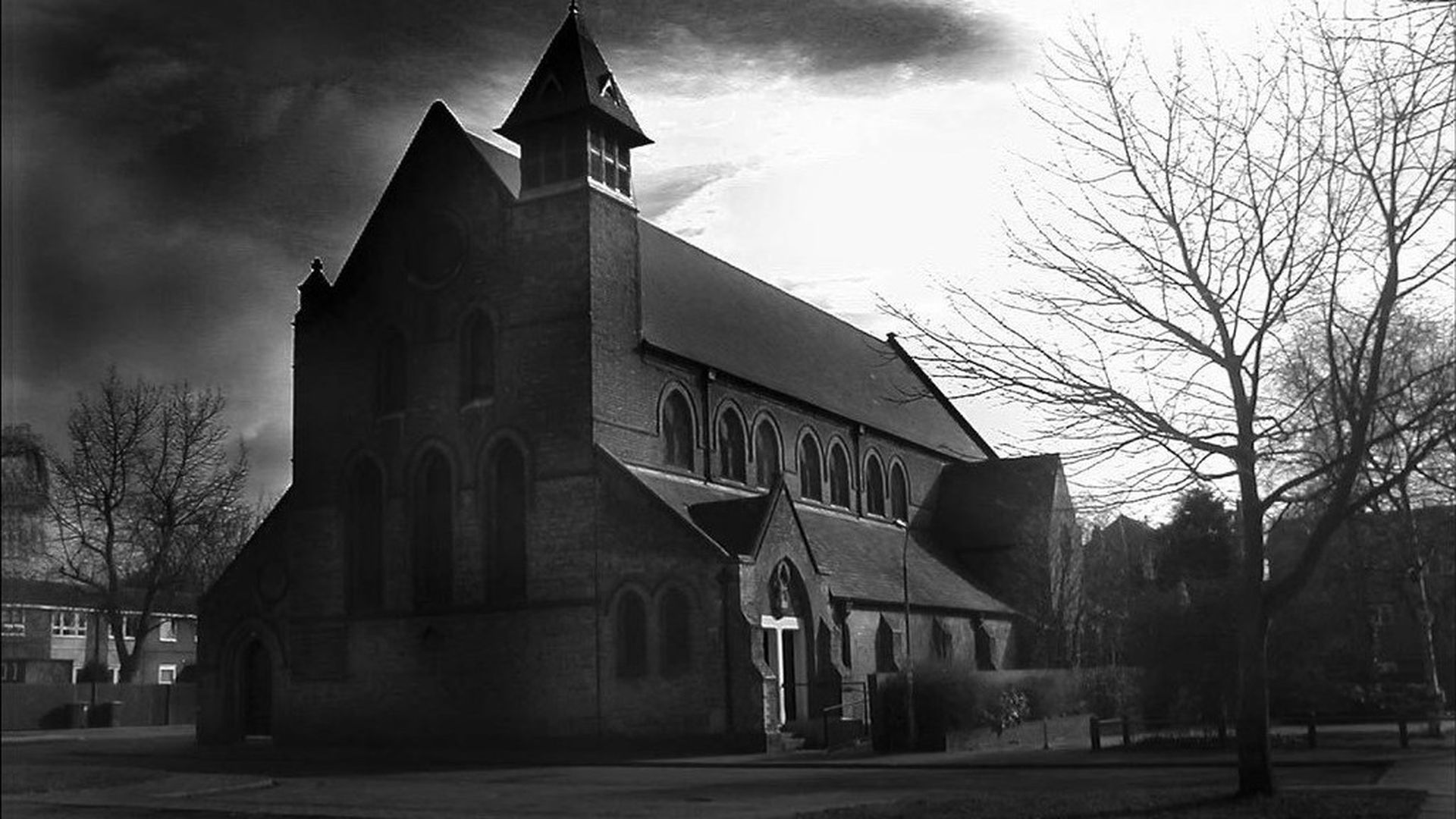 12. El oscurecimiento de la Iglesia. Black and white image of a Gothic-style church under a dramatically cloudy sky, with leafless trees to the right and a crucifix visible near the entrance. Opens a video in a new window.