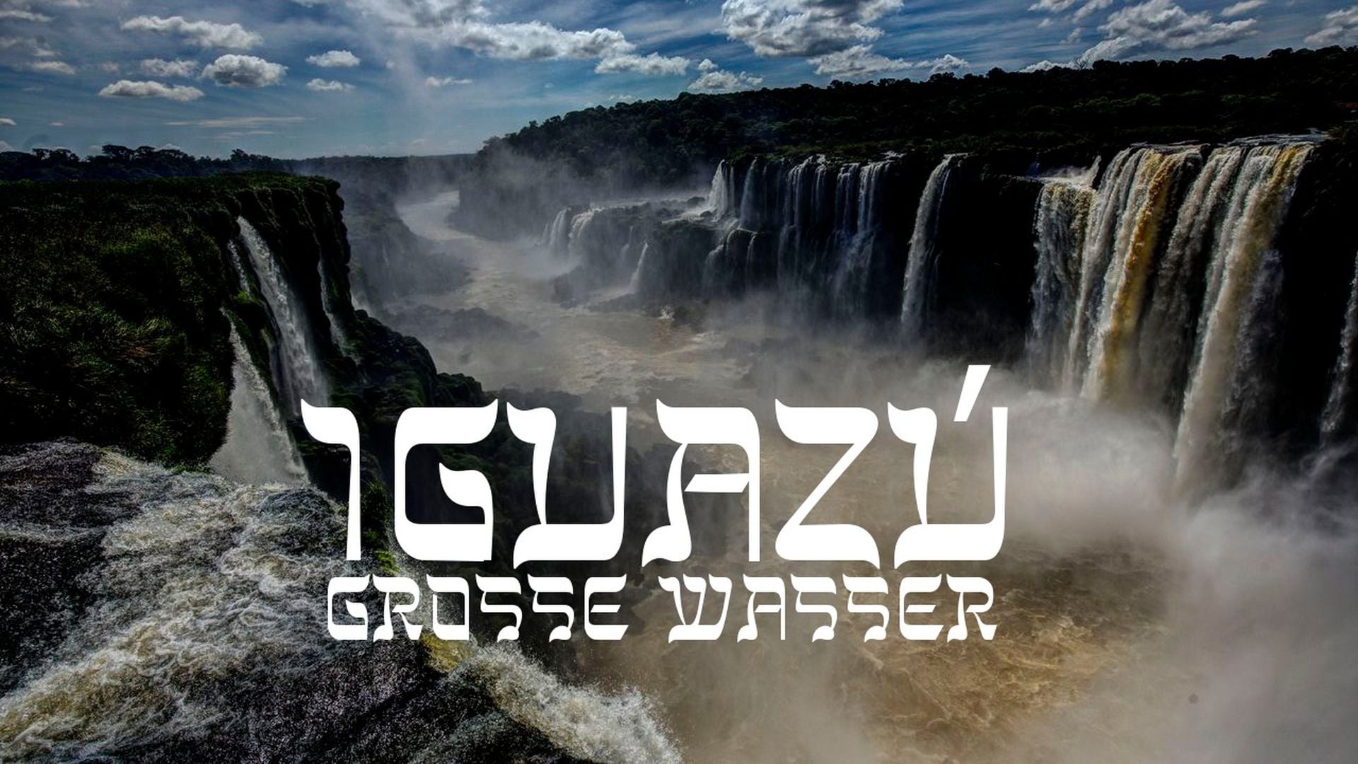 19. Iguazú - Große Wasser. Aerial view of the majestic Iguazu Falls under a bright sky, with powerful water cascades generating mist. The image includes text overlays, "IGUAZÜ" at the top in large letters and "GROSSE WASSER" at the bottom in smaller letters, which translate to 'great water' in German. Opens a video in a new window.