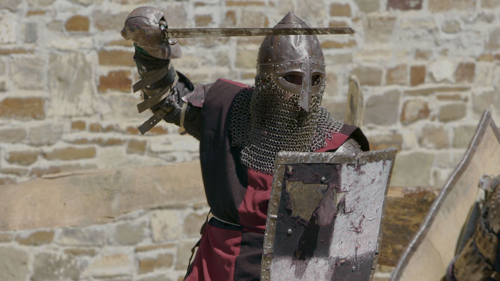 2. El grito de guerra del enemigo. A knight in traditional armor, complete with a chainmail hood and helmet, holds a sword in a striking posture behind a weathered shield, against a backdrop of an ancient stone wall. Opens a video in a new window.