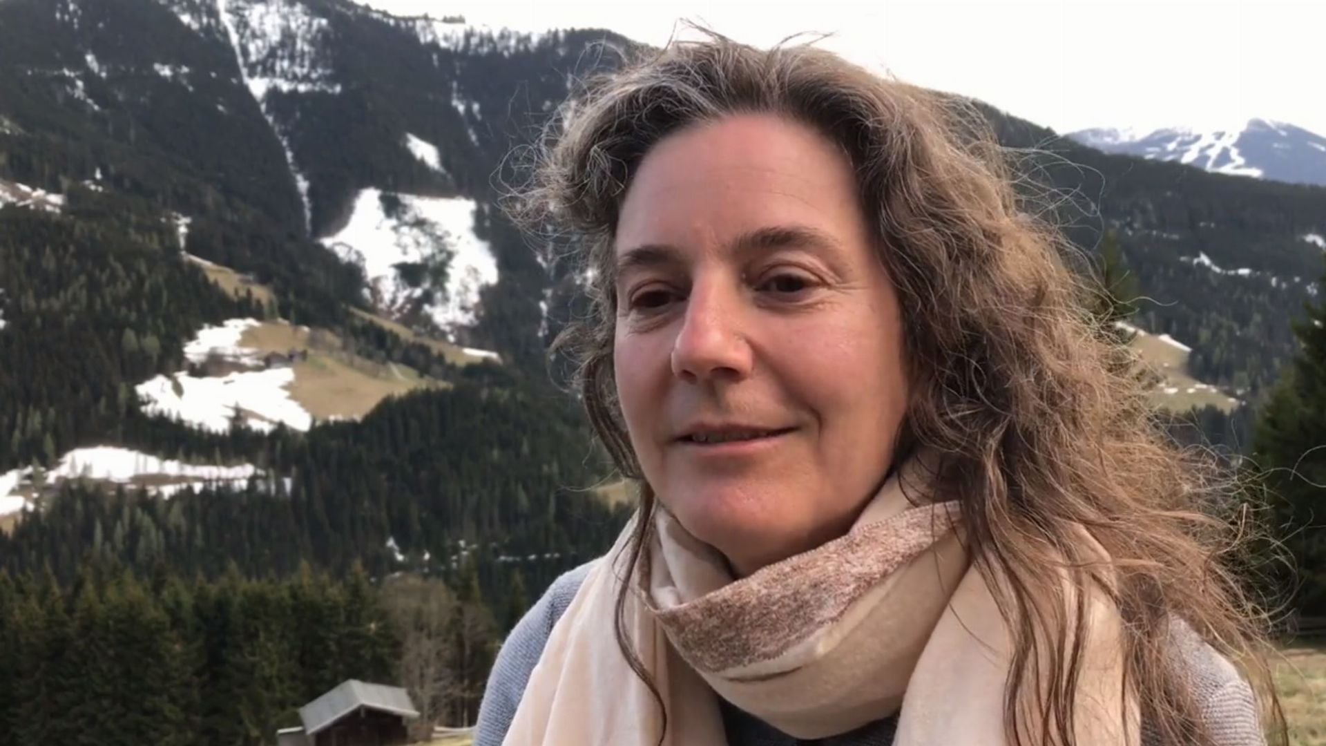 18. Unverdient. A woman with curly hair, wearing a light scarf, smiles gently in the foreground. Behind her, a scenic mountain landscape with patches of snow and coniferous trees under a cloudy sky unfolds. Opens a video in a new window.