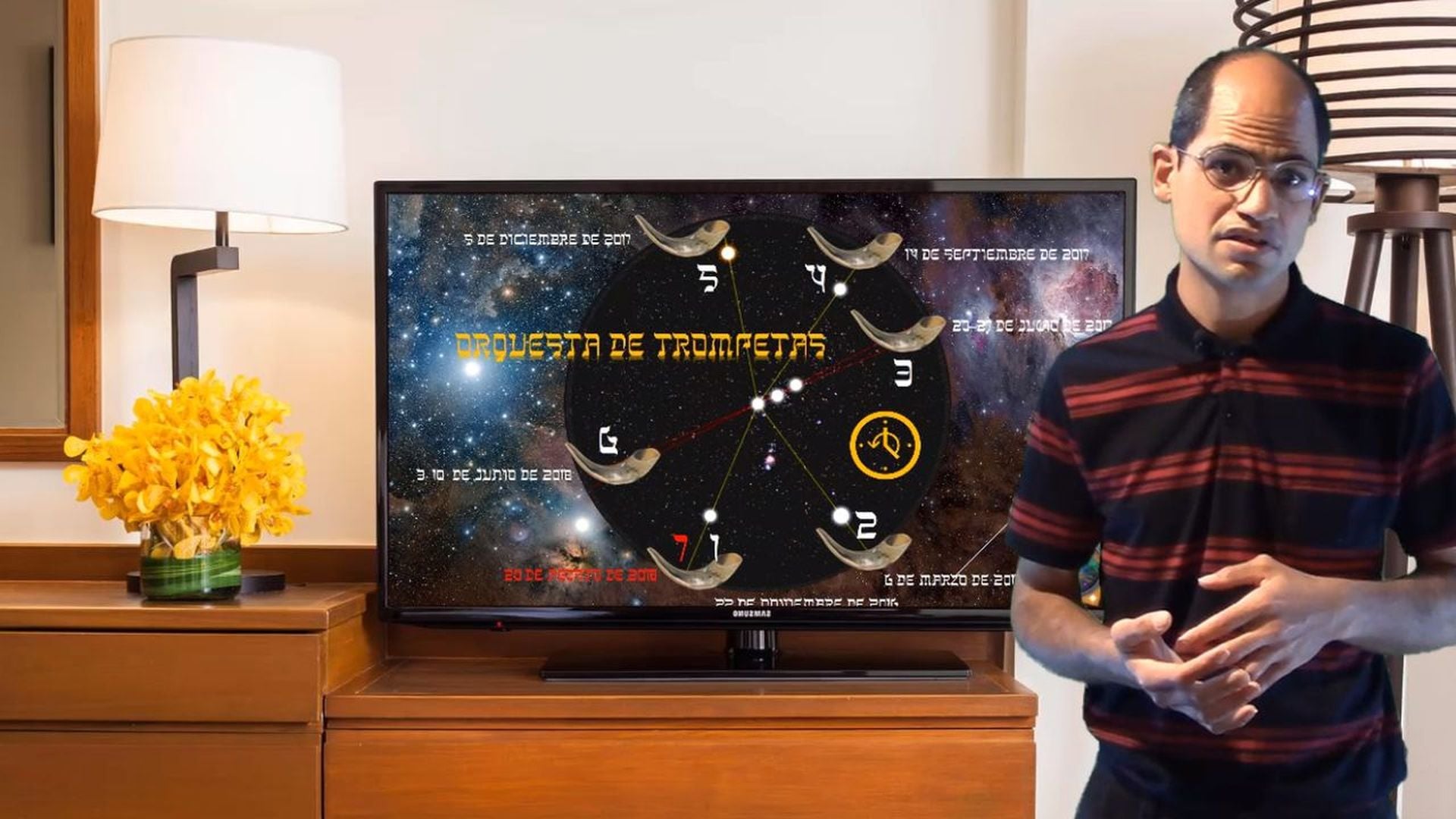 5. ¡Prepárate para los ayes!. A man with glasses, balding and wearing a red striped shirt, stands next to a TV displaying a graphical representation of celestial constellations as outlined in the biblical Mazzaroth. The TV is on a wooden console with a white lamp and a vase of yellow flowers beside it. Opens a video in a new window.