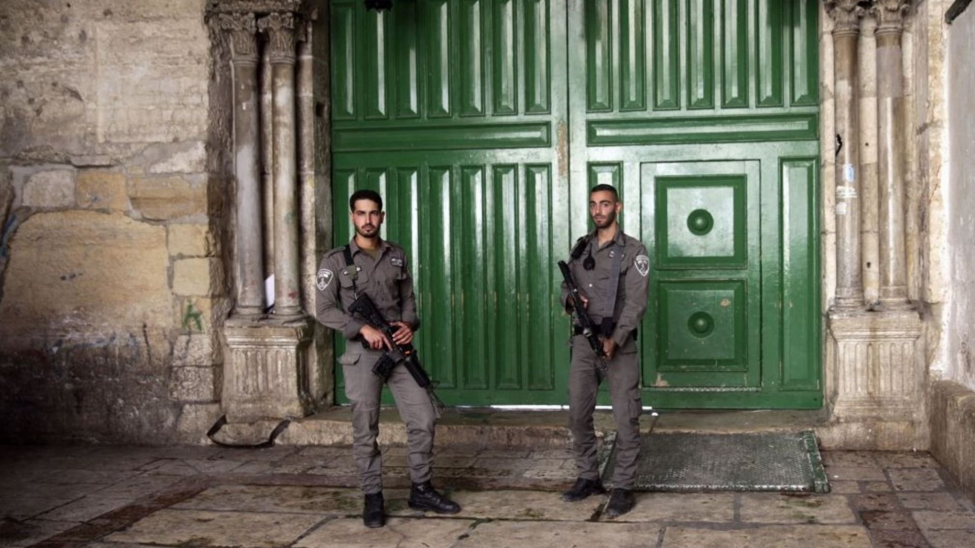 2. Israel advierte que los detectores de metal seguirán en entradas de la mezquita de Al Aqsa. Two security personnel in uniform stand guard in front of a large green door set within an ancient stone wall, the architecture suggesting a historical significance under the night sky. Opens a video in a new window.