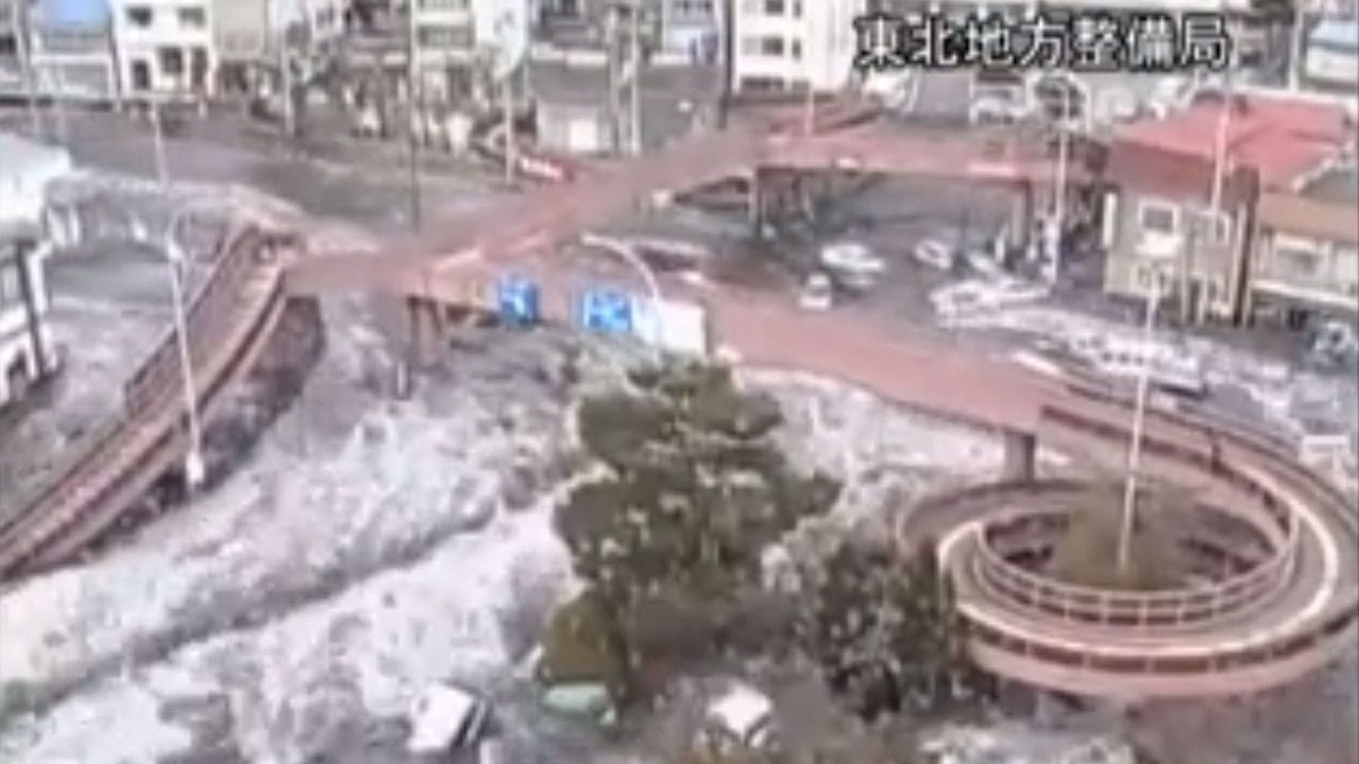 8. 2011 Japan Tsunami Caught on CCTV Cameras. Aerial view of an urban area with roads and buildings partially covered by snow, alongside a clear view of a circular structure and green trees. Opens a video in a new window.