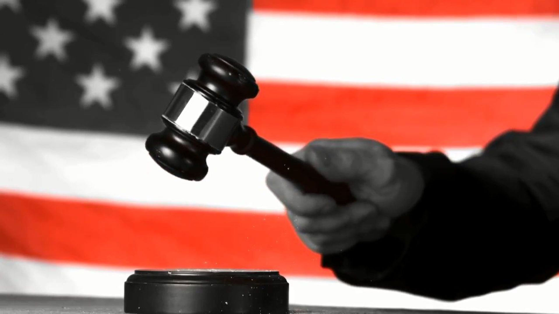 12. LAW MANDATORY SUNDAY WORSHIP!. A monochrome image selectively colored depicting a hand striking a gavel down, set against a blurred backdrop of the American flag. Opens a video in a new window.