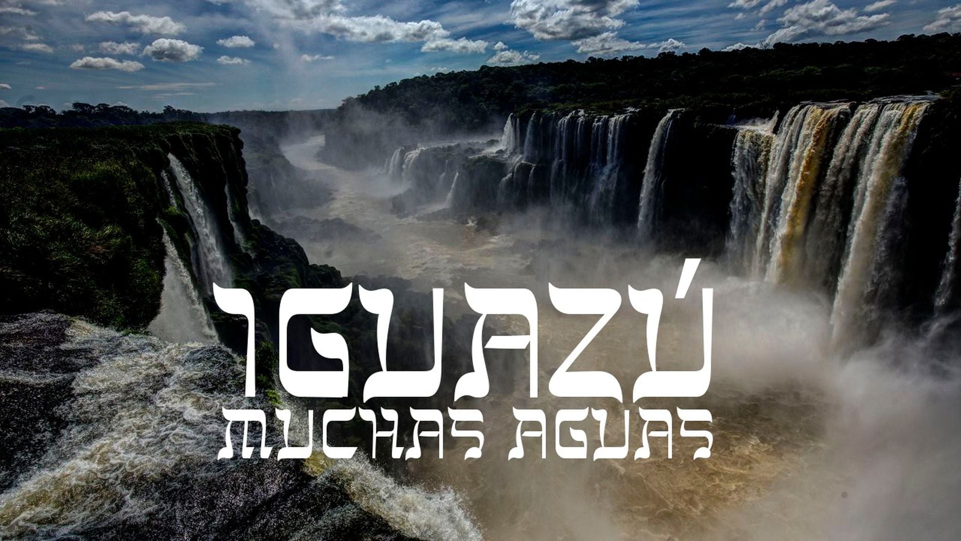 22. Iguazú - Muchas Aguas. Panoramic view of the majestic Iguazú Falls with numerous cascades spilling vast amounts of water, surrounded by lush greenery under a partly cloudy sky. Overlay text reads "IGUAZÚ MUCHAS AGUAS" in bold, stylized font. Opens a video in a new window.