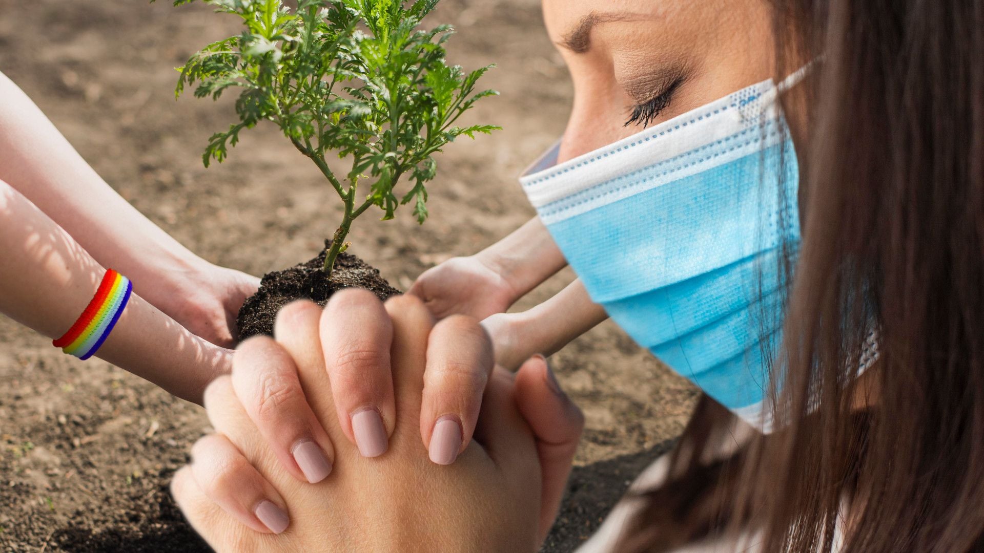 16. Das dreifaltige Malzeichen des Tieres. A young woman wearing a surgical mask and two other individuals plant a small tree together in soil. The woman's eyes are closed, and one of the hands assisting her displays a rainbow-colored bracelet. Opens a video in a new window.
