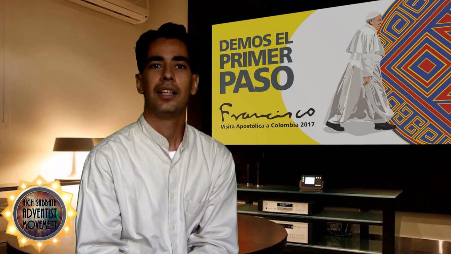 19. Pope in Colombia, Sept. 9—Deep Meaning!. A man in a white shirt is seated in front of a poster displaying a graphical illustration depicting a figure in white robes and a slogan related to a 2017 event in Colombia. A small digital clock sits on a desk beside him, as well as a logo related to a movement features prominently in the bottom left of the image. Opens a video in a new window.