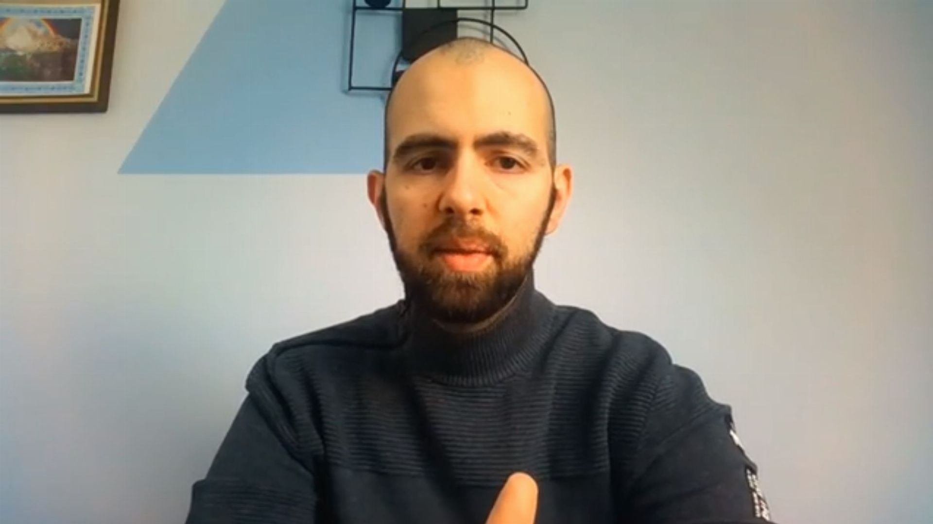 17. Ich habe Gott geleugnet - und Er schenkte mir Gnade. Man with a beard in a dark turtleneck sits in front of a camera with a serene expression. A light blue wall adorned with a minimalistic metal frame artwork is behind him. Opens a video in a new window.