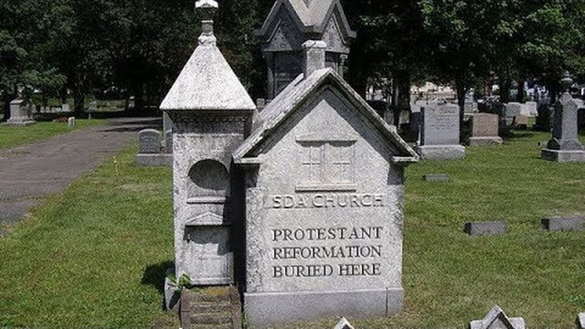 5. SDA Church, the tombstone of the Reformation. A pair of ornate, small stone mausoleum structures in a cemetery, one marked "SDA Church" and the other labeled "Protestant Reformation Buried Here." Surrounded by numerous other gravestones under a clear sky. Opens a video in a new window.
