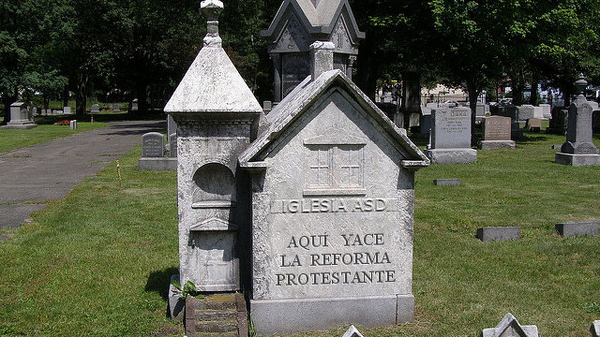 10. Iglesia ASD, la lápida de la Reforma. A unique stone monument in a cemetery, resembling a small church, with the inscription "Iglesia ASD Aqui Yace La Reforma Protestante" under a cross, surrounded by various other gravestones under a clear sky. Opens a video in a new window.