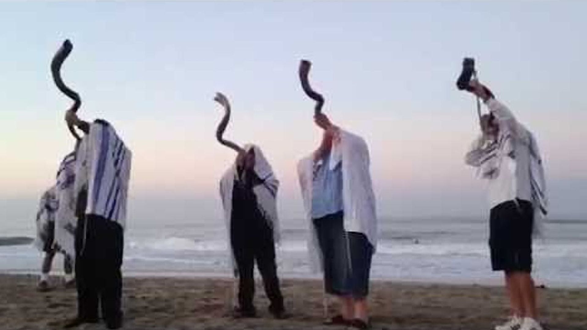 1. Shofars durante los vigilantes del crepúsculo. Four individuals on a beach at dusk, each holding a shofar, a traditional horn used in ceremonies, to their mouths. They wear prayer shawls over their heads, facing the sea as waves gently lap the shore. Opens a video in a new window.