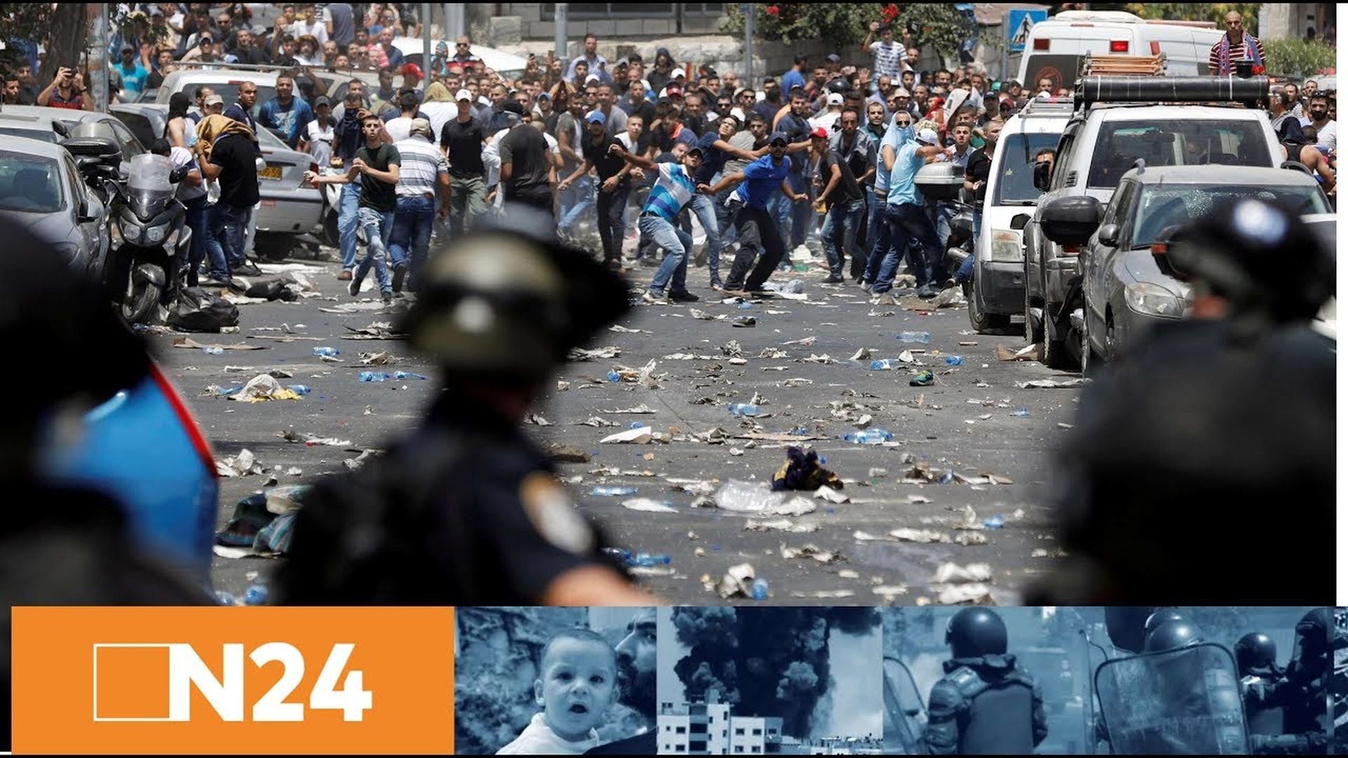 2. Tempelberg-Krise eskaliert: Tote und Verletzte - Palästinenser brechen Kontakte zu Israel ab. A chaotic scene on a city street with numerous individuals, many in civilian clothes, clashing among scattered debris, while law enforcement officials in riot gear observe and manage the situation. The foreground focus is on a police officer observing the turmoil. Opens a video in a new window.