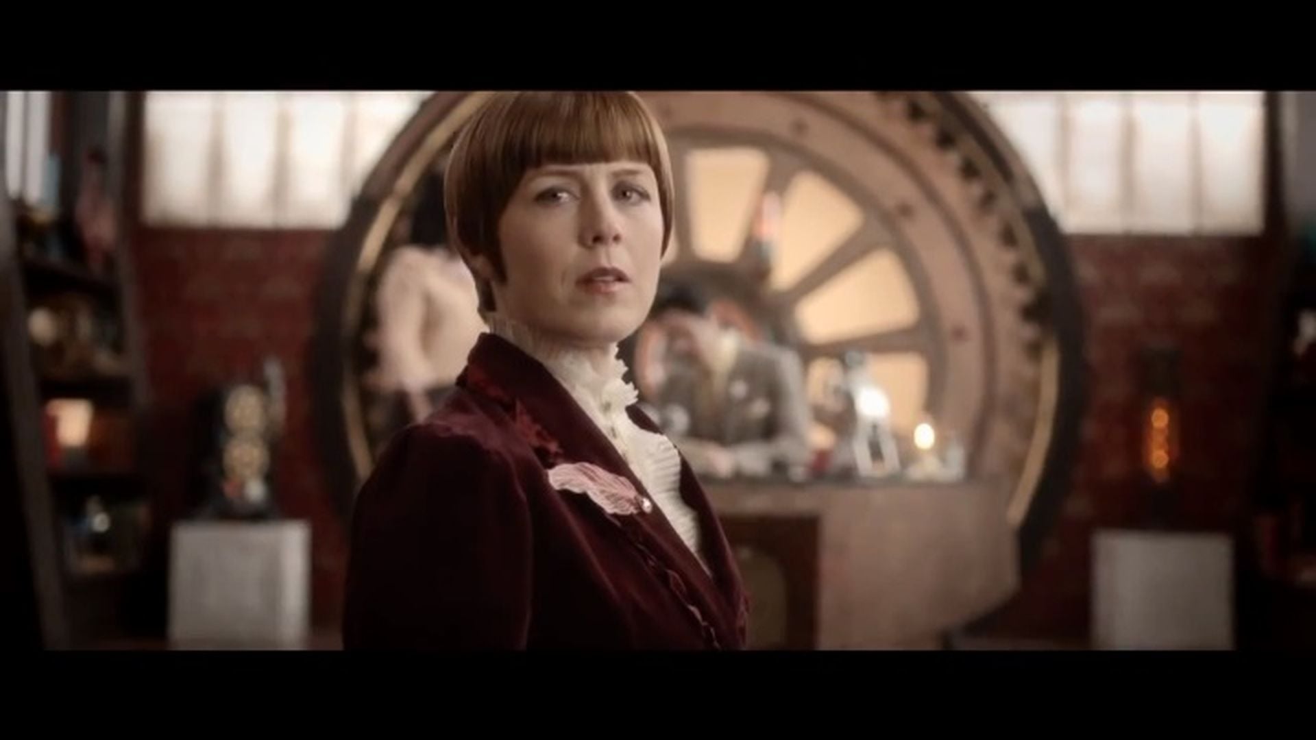 1. Der Schriftwart – Offizieller Trailer. A woman with a short brown bob haircut and a burgundy blazer stands confidently in an ornate room filled with antique furniture and circular, metallic artifacts resembling astronomical instruments around the walls. The background gives a classical yet mystical ambiance reminiscent of a study dedicated to the Mazzaroth. Opens a video in a new window.