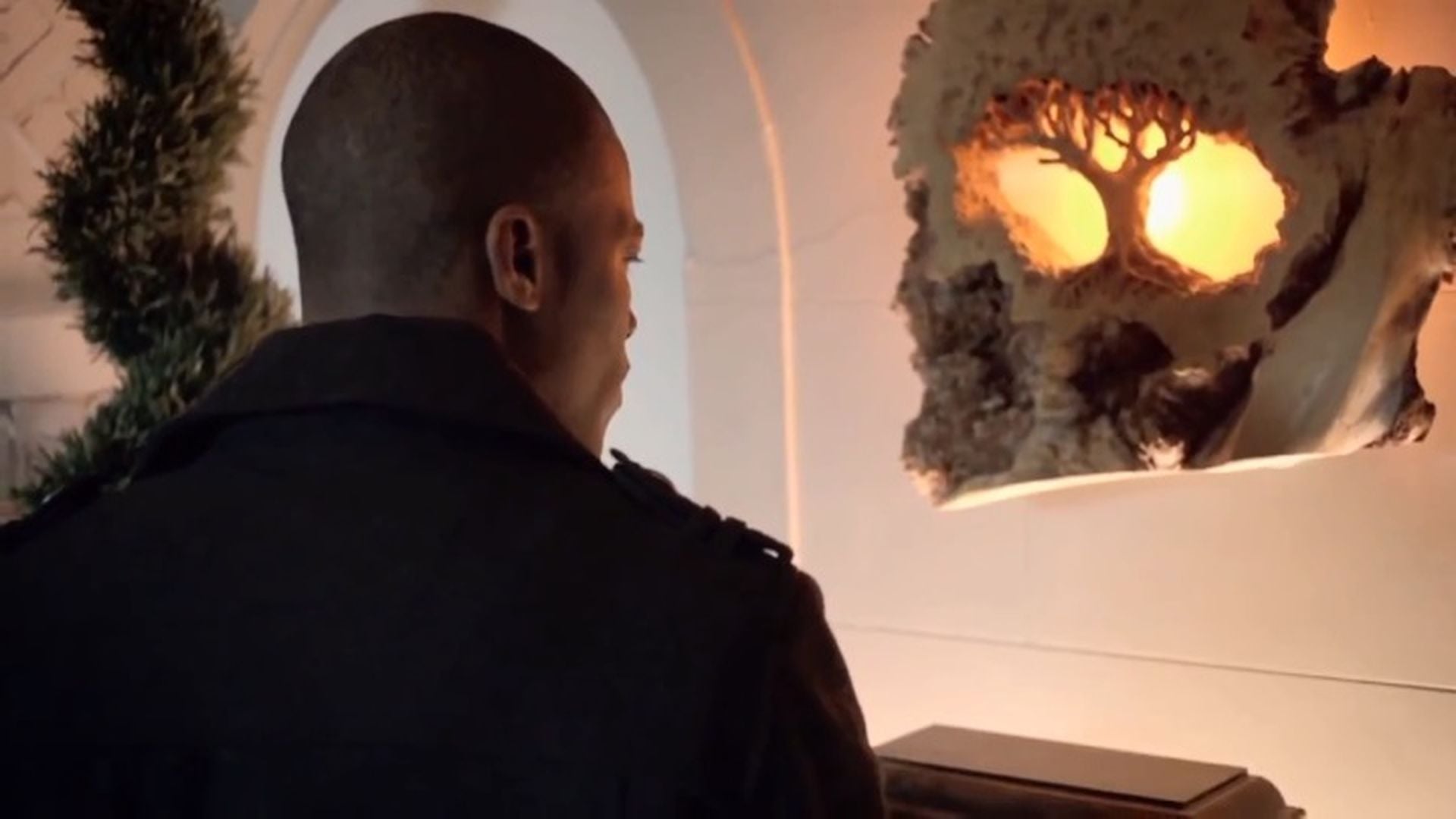 2. Der Schriftwart - Pilotfolge. A man with a shaved head, viewed from behind, gazes at an illuminated sculptural relief depicting a celestial theme reminiscent of the Mazzaroth, mounted on a white arched wall. The artwork radiates a warm, golden glow, and a subtle wreath decorates the arched entry. Opens a video in a new window.