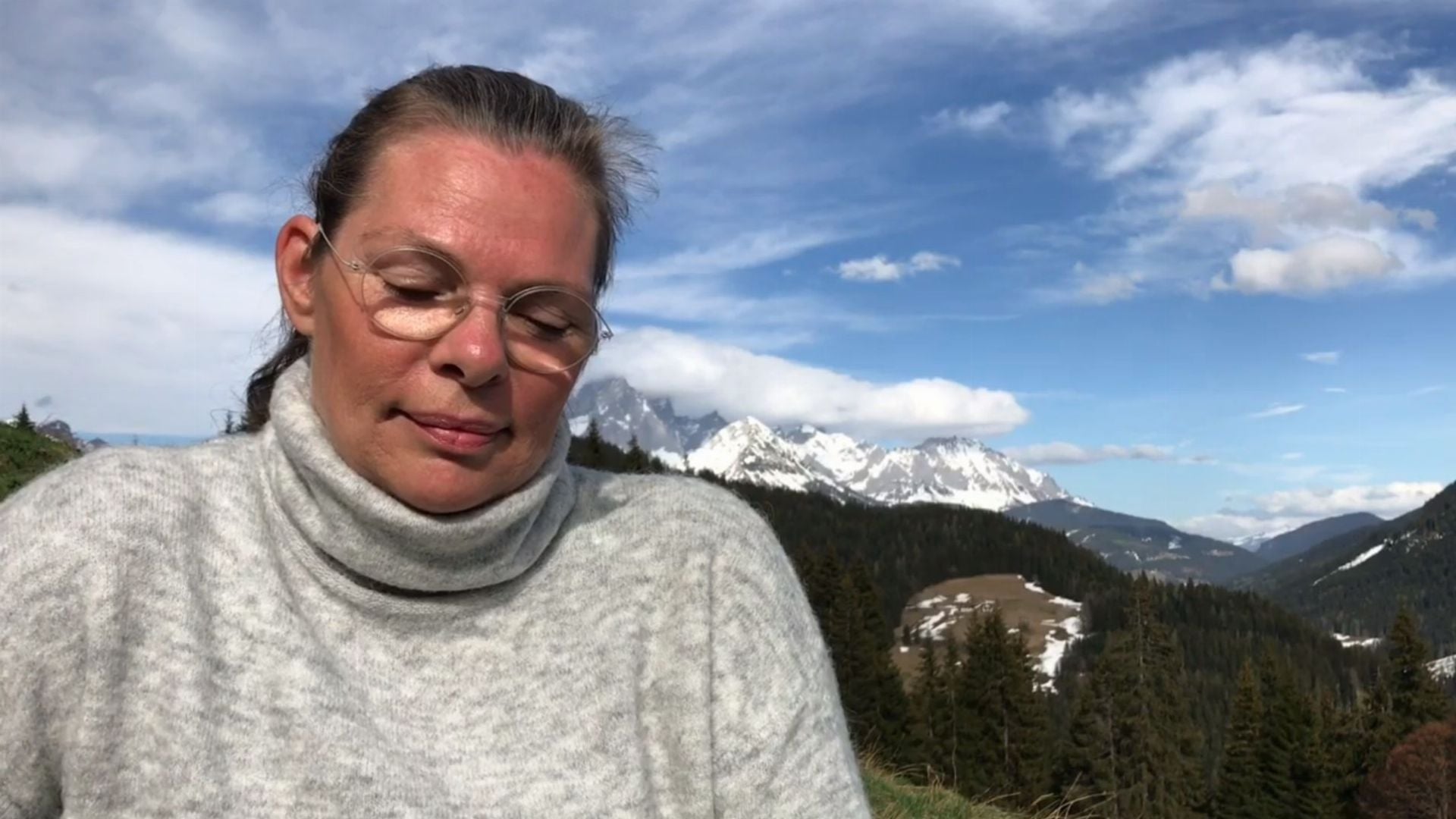 19. Jesus ist die Liebe. A woman with her eyes closed, wearing a light grey turtleneck sweater, stands in the foreground against a majestic mountain landscape under a clear blue sky. Opens a video in a new window.