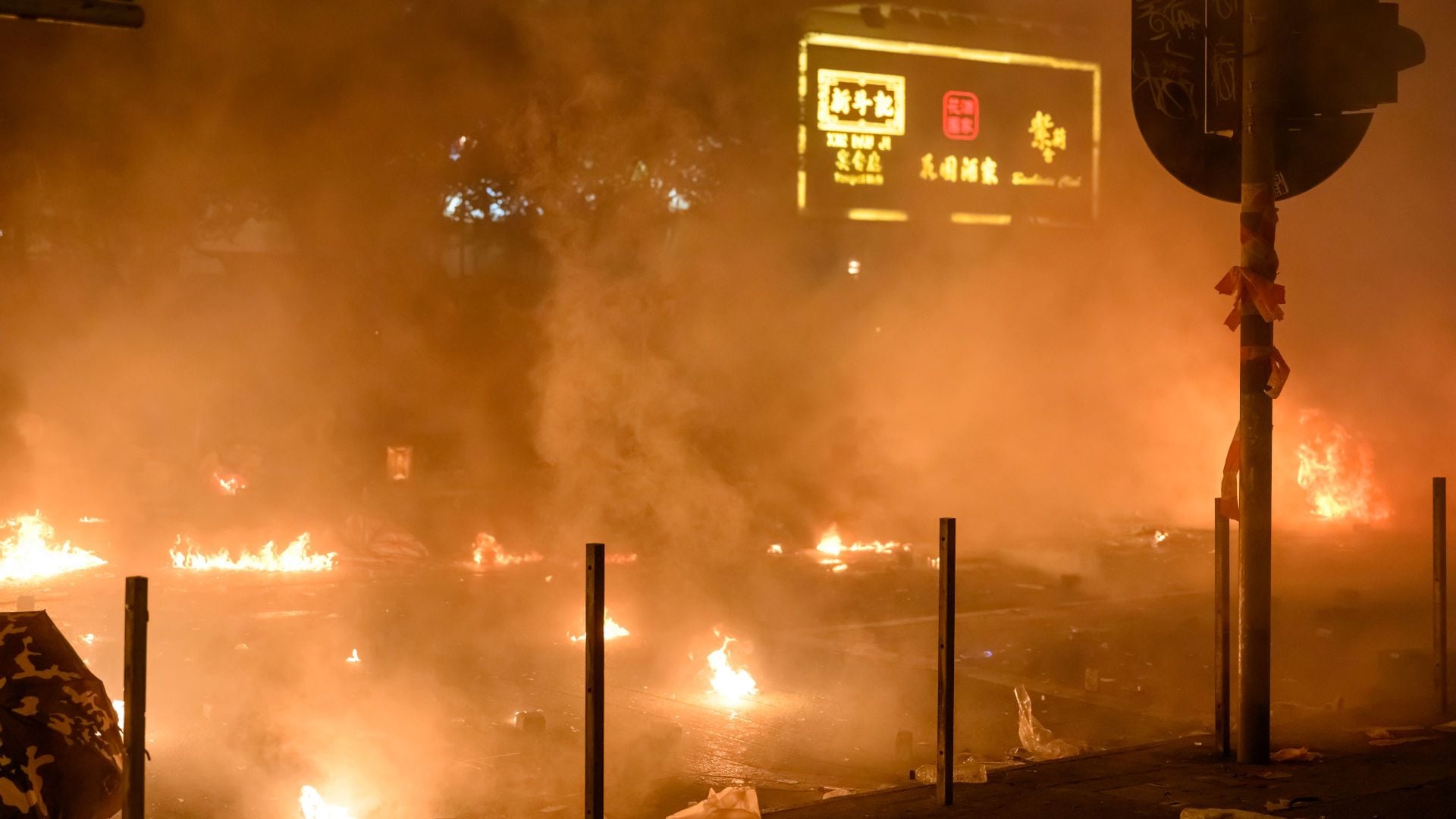 9. Fuego en el Monte Carmelo. A nocturnal city scene illuminated by multiple small fires scattered across a street. Smoke obscures parts of the background, which includes an illuminated street sign displaying characters in a non-Latin script. Opens a video in a new window.