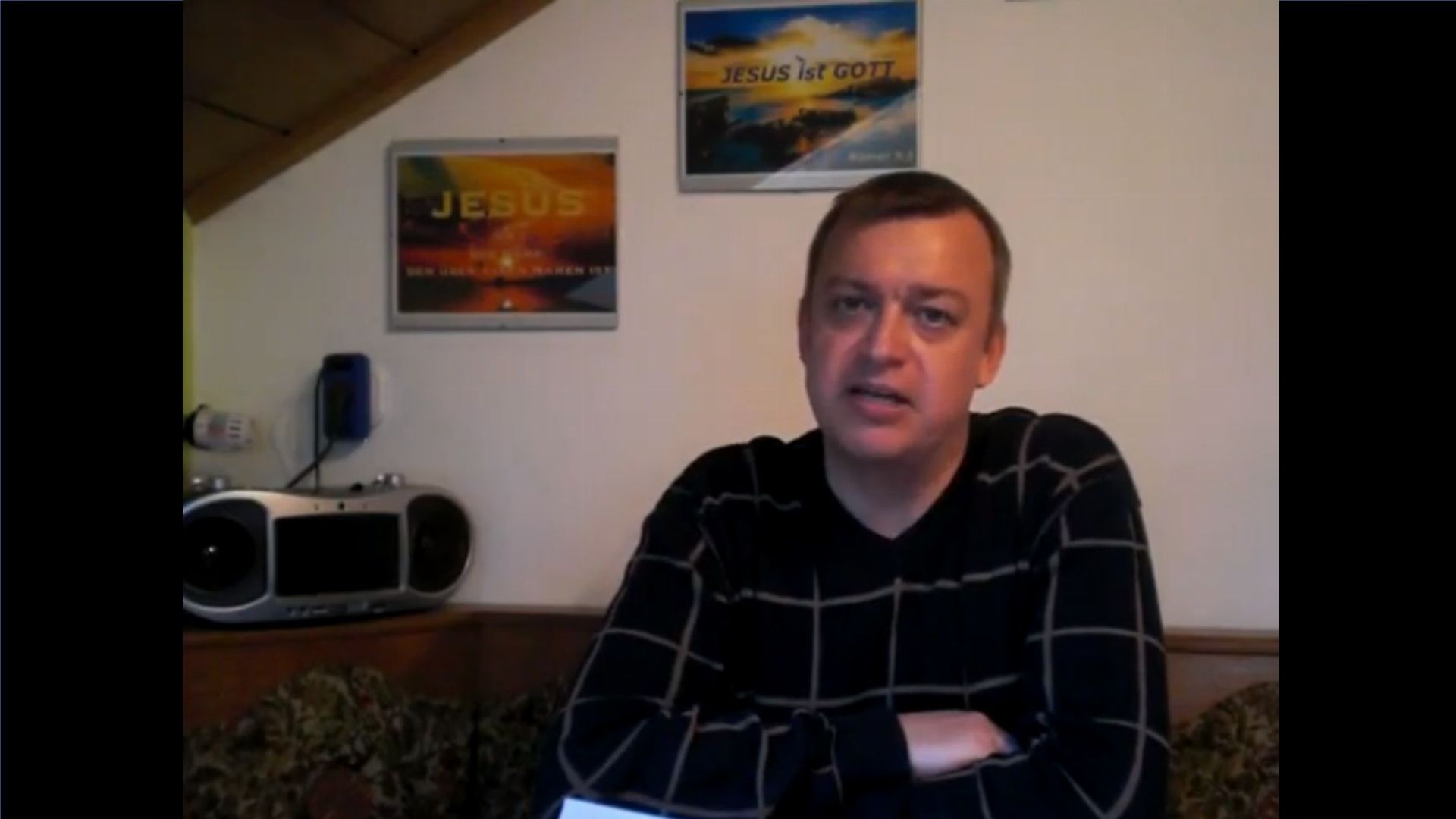 10. Der Glaube Jesu. A man in a black and gray sweater is seated indoors, speaking. The room features a rustic decor with religious-themed posters saying "Jesus is GOD" and a depiction of a sunset. A stereo system rests on the side. Opens a video in a new window.