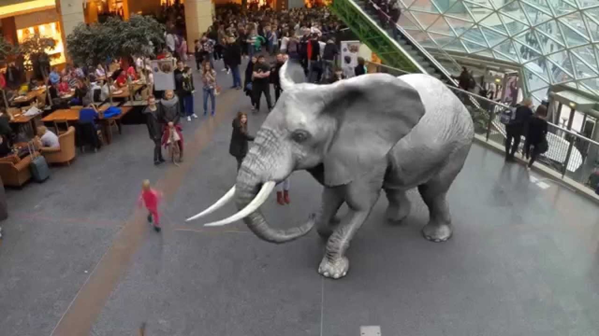 7. Zoo der Erweiterten Realität. A digitally imposed elephant stands in the center of a bustling shopping mall with multiple levels and a glass ceiling. People of various ages are scattered around the mall, some walking and others seated at dining tables. Opens a video in a new window.