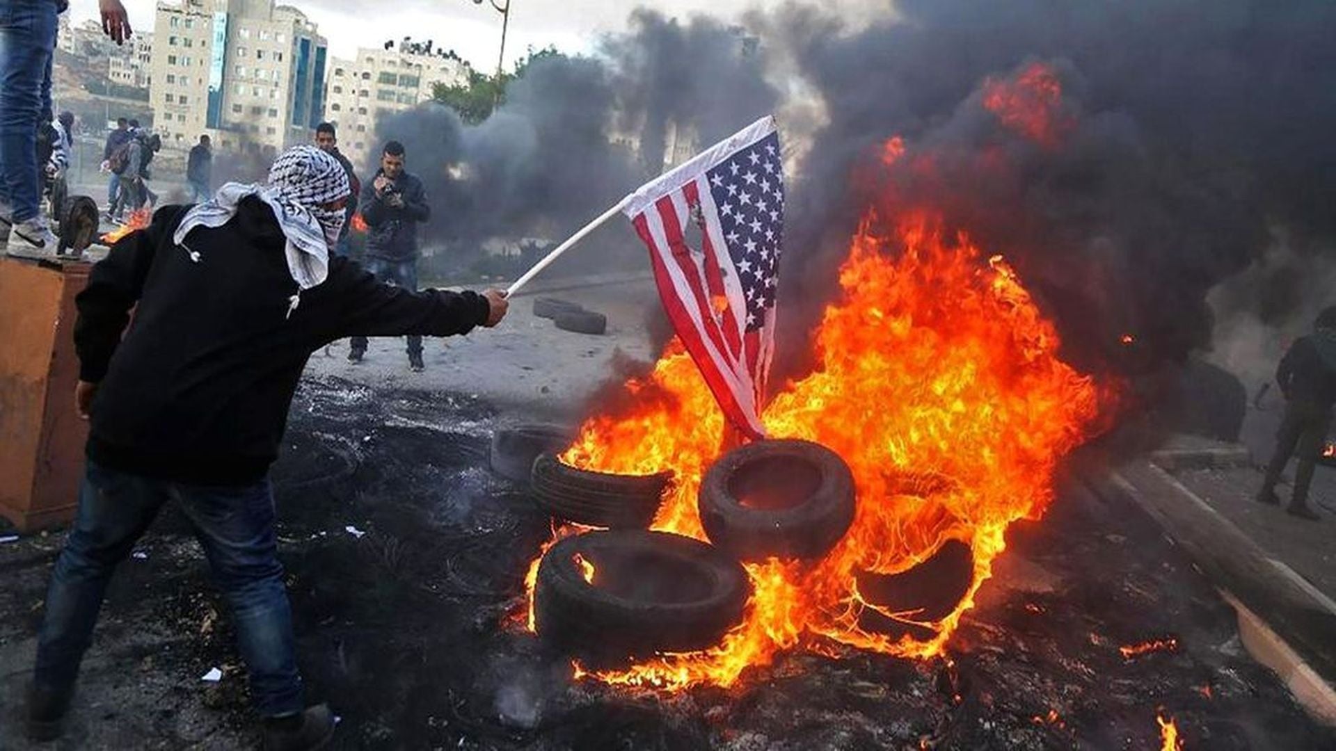 4. Israel Heats Up the Great Furnace. A person with a covered face holding an American flag over a fire made from burning tires in an urban setting with buildings visible in the background. Smoke fills the air. Opens a video in a new window.