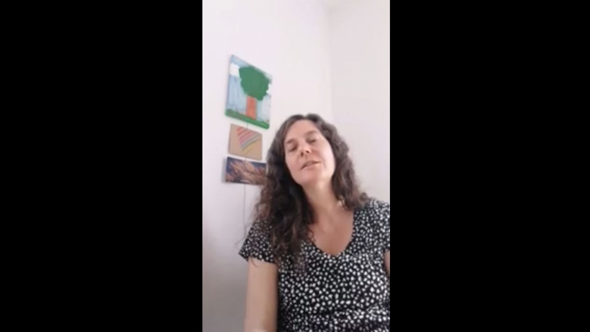 1. Jesus kommt!. A woman with curly hair sits smiling softly in a casually dressed room. Behind her, a simplistic painting resembling terrestrial topography hangs on a white wall. Opens a video in a new window.