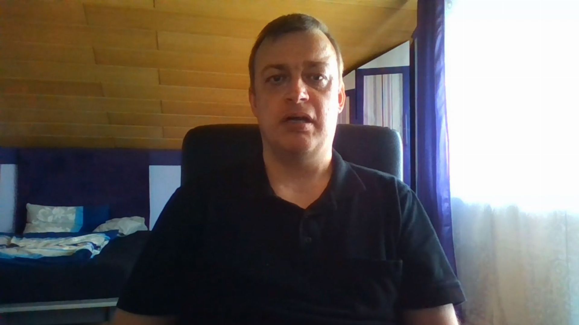 21. Herzenswunsch. A man seated in a wood-paneled room with a bed in the background. He wears a black shirt and appears to be speaking. Opens a video in a new window.