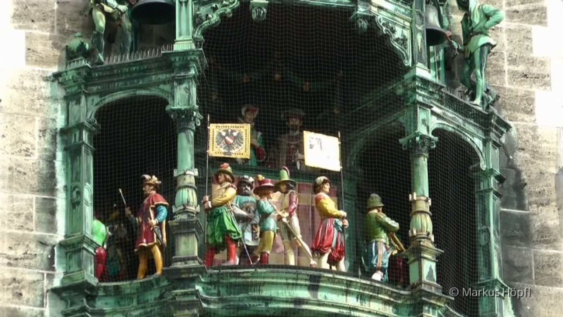 6. Carillon on Mary's Square in Munich. Mechanical figures in historical costumes perform on an ornate clock tower's balcony, surrounded by sculptural details and stone architecture. The central figure holding a shield adorned with a starry representation, possibly referring to the Mazzaroth, is flanked by others with musical instruments and regal attire. Opens a video in a new window.