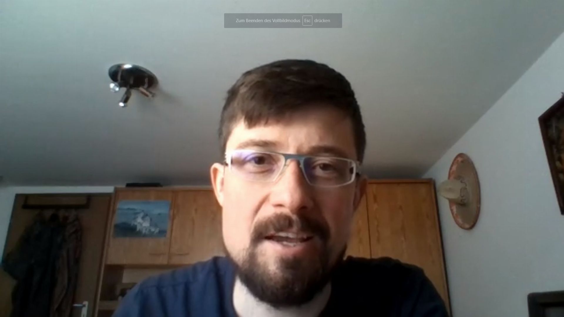 13. Was für eine wunderbare Veränderung in meinem Leben. A man with eyeglasses and a beard is participating in a video call, presenting a homely background that includes a wall-mounted light fixture and a framed picture of a maritime scene. Opens a video in a new window.