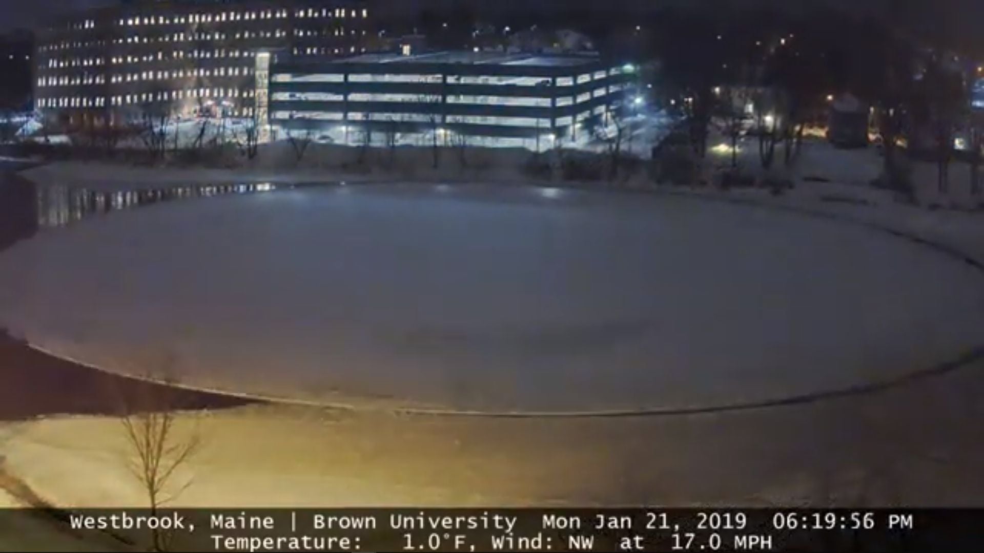 14. Westbrook-Eisscheibe - Zeitraffer vom 21. Januar 2019. A winter night scene showing a frozen pond in the foreground with a softly lit multi-story building in the background. The sky appears clear, reflecting a typical cold evening under a starry firmament. The image includes a timestamp and weather data: Westbrook, Maine, Brown University, with a temperature of 1.0°F and a northwest wind at 17.0 MPH, dated January 21, 2019 at 06:19:56 PM. Opens a video in a new window.