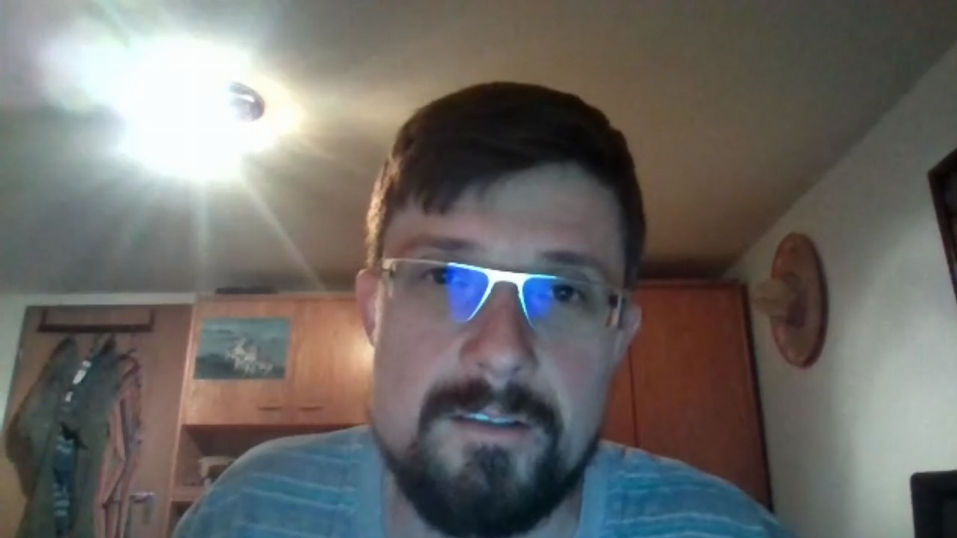 15. Die vierte Engelsbotschaft. A man with a beard and glasses sitting indoors with a bright light behind his head resembling a star in the celestial firmament, in an environment with wooden furniture and items hanging on the wall behind him. Opens a video in a new window.