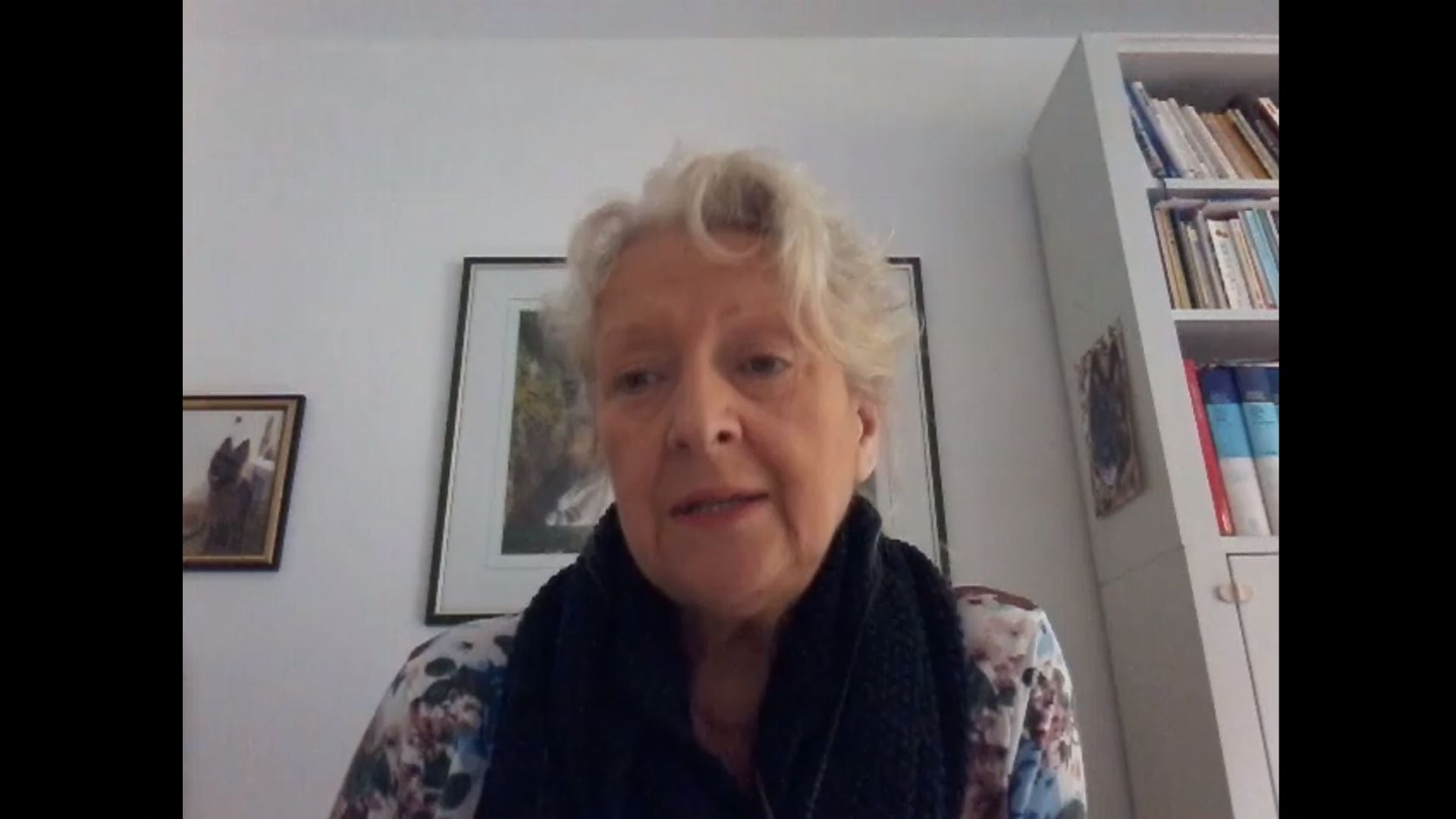 24. Die Liebe Jesu. An elderly woman with light hair wearing a scarf and a floral blouse speaks during a video call. A bookshelf filled with books and a wall decorated with framed pictures are visible in the background. Opens a video in a new window.