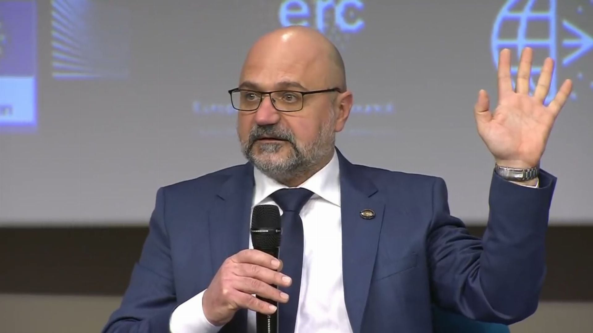 1. LIVE: Astronomen geben bahnbrechende Ergebnisse von Schwarzlochbeobachtungen bekannt.. A man with a beard dressed in a suit holds a microphone and gestures with his left hand while speaking at a conference. The background displays a logo with a globe and the initials "ERC." Opens a video in a new window.